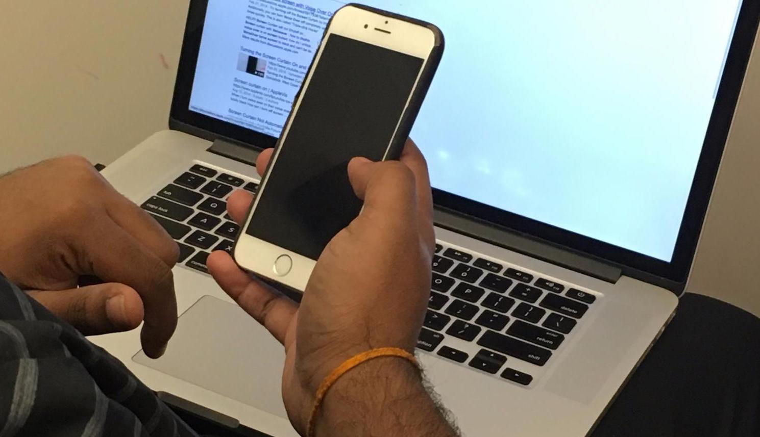 A male hand is holding an iPhone with a black screen in front of an open laptop.